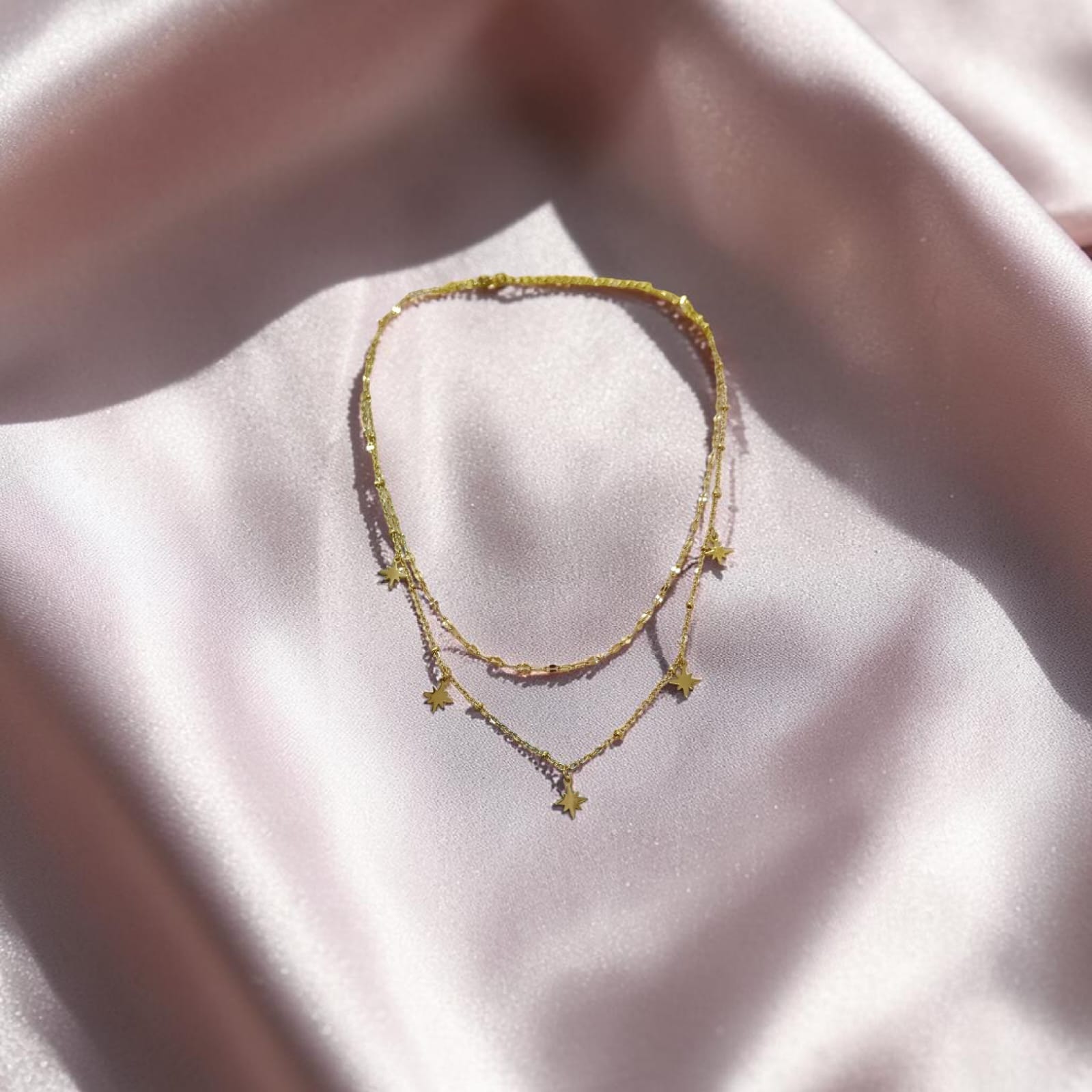 A delicate double-strand gold chain necklace with small star charms, displayed on a soft, draped light pink satin fabric.