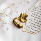 A polished gold-toned abstract sculptural ring or brooch with a glossy finish, styled on a white fabric surface next to a torn vintage book page and small white flowers.
Grishita Jewellery