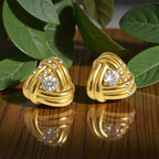 A pair of gold knotted earrings with clear round gemstones and small clear accent stones, displayed on a reflective wooden surface with green leaves in the background.