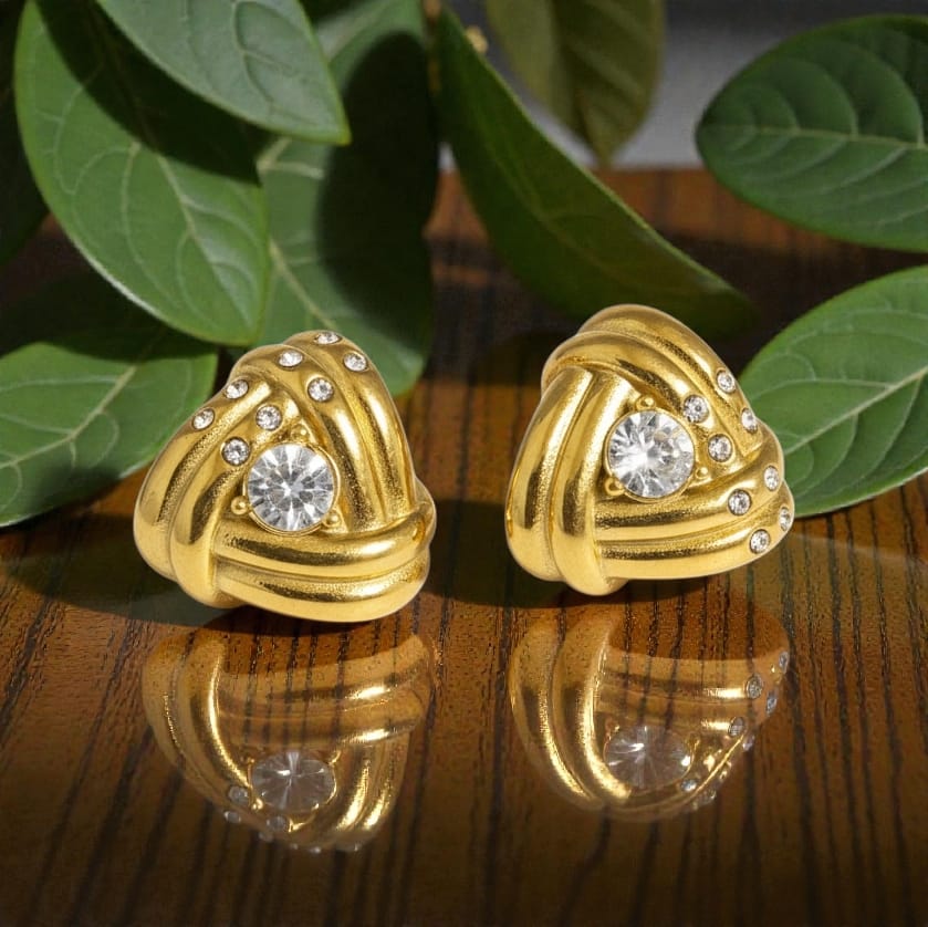 A pair of gold knotted earrings with clear round gemstones and small clear accent stones, displayed on a reflective wooden surface with green leaves in the background.